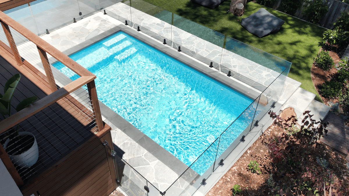 A stylish rectangular swimming pool with glass fencing and surrounding garden.