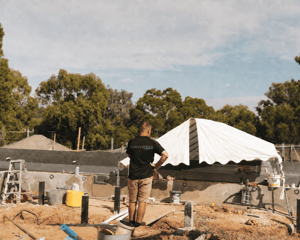 Construction worker at a pool site with a white tent and trees in the background.